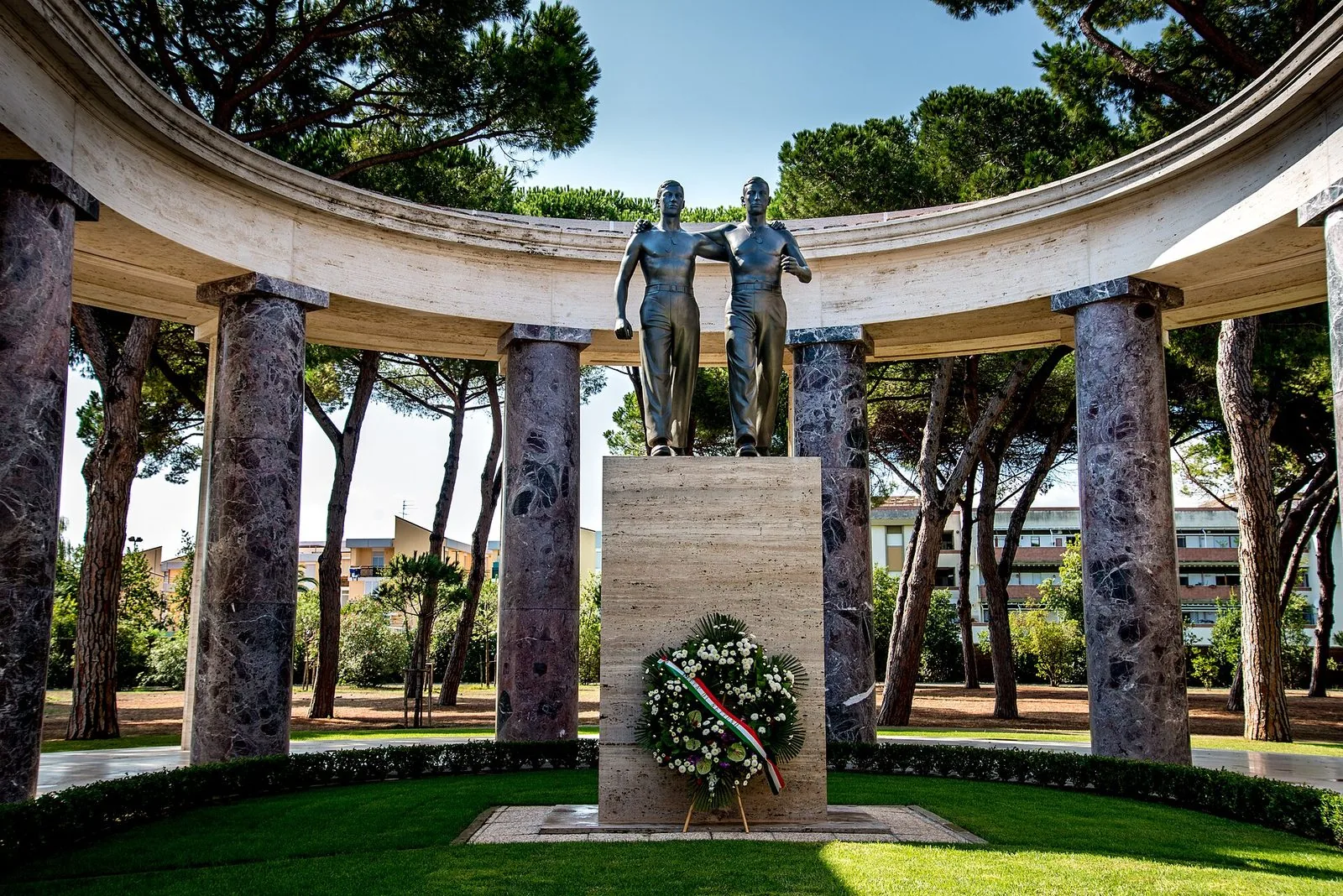 Il monumento dei due soldati abbracciati al Cimitero americano di Nettuno, una scultura in bronzo su un basamento di travertino con una corona di fiori bianchi e nastro tricolore, al centro di un colonnato semicircolare fra i pini