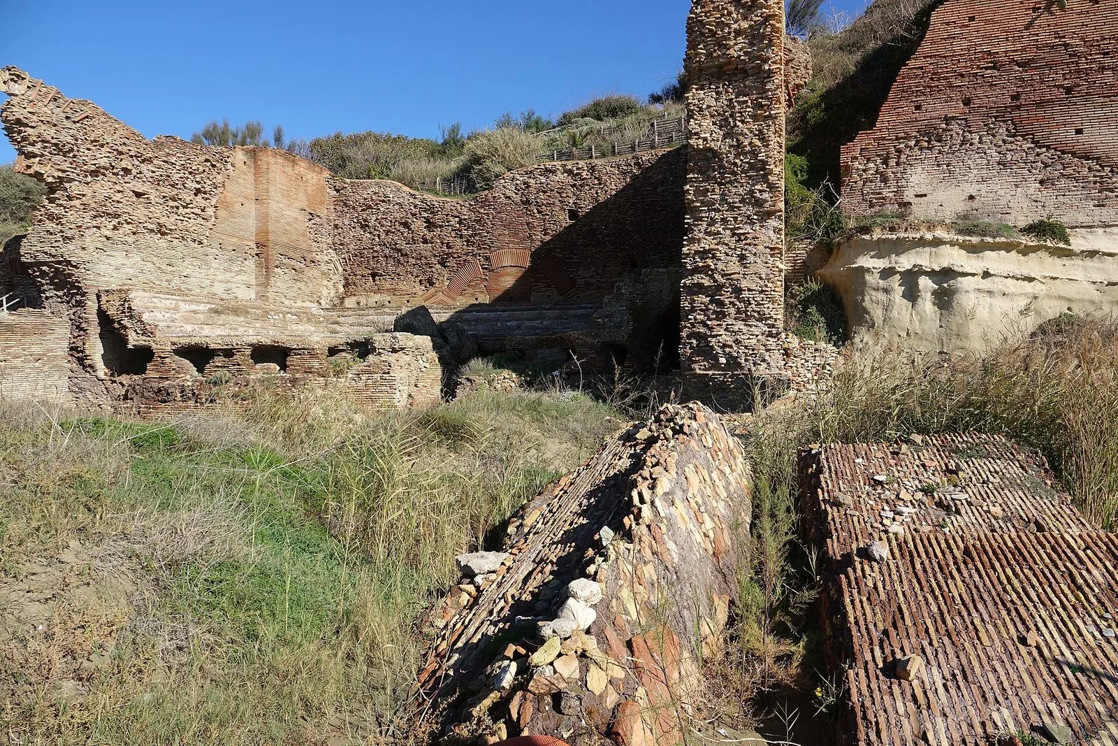 Rovine in laterizio della Villa di Nerone ad Anzio: muri romani a strati di mattoni con un'abside decorata, un pilastro e frammenti crollati fra l'erba alta, sotto la falesia chiara e un cielo azzurro