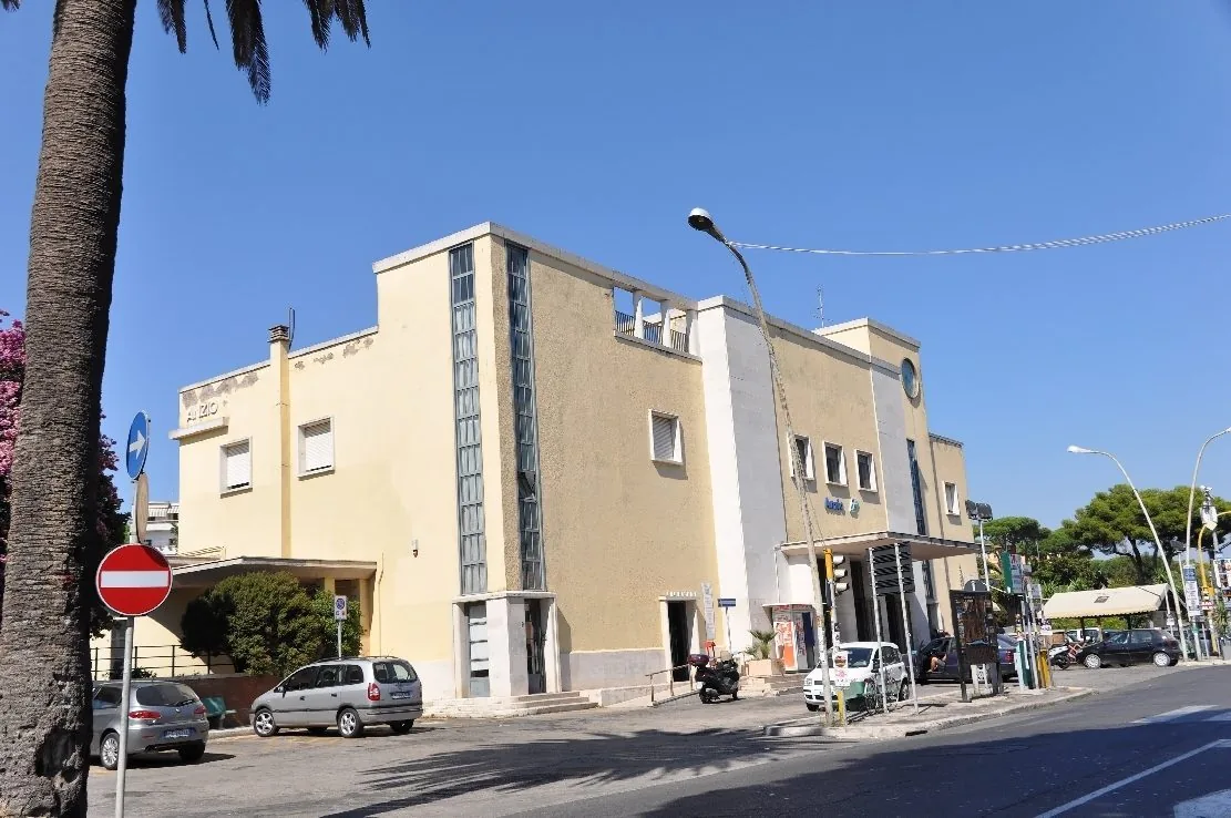 L'edificio della stazione ferroviaria di Anzio, con la facciata color crema, le finestre a nastro in vetro e la scritta Anzio, sotto un cielo azzurro con auto parcheggiate davanti