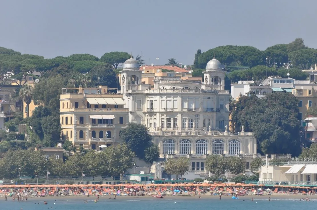 Il Paradiso sul Mare di Anzio, edificio Liberty bianco con due cupolette e grandi finestre ad arco, affacciato su una spiaggia con file di ombrelloni arancioni e circondato da pini e palazzi color sabbia