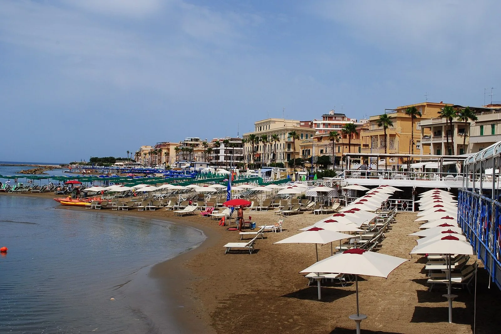 Una spiaggia di sabbia ad Anzio d'estate, con file di ombrelloni bianchi e lettini, l'acqua calma a sinistra e i palazzi chiari con le palme lungo il lungomare
