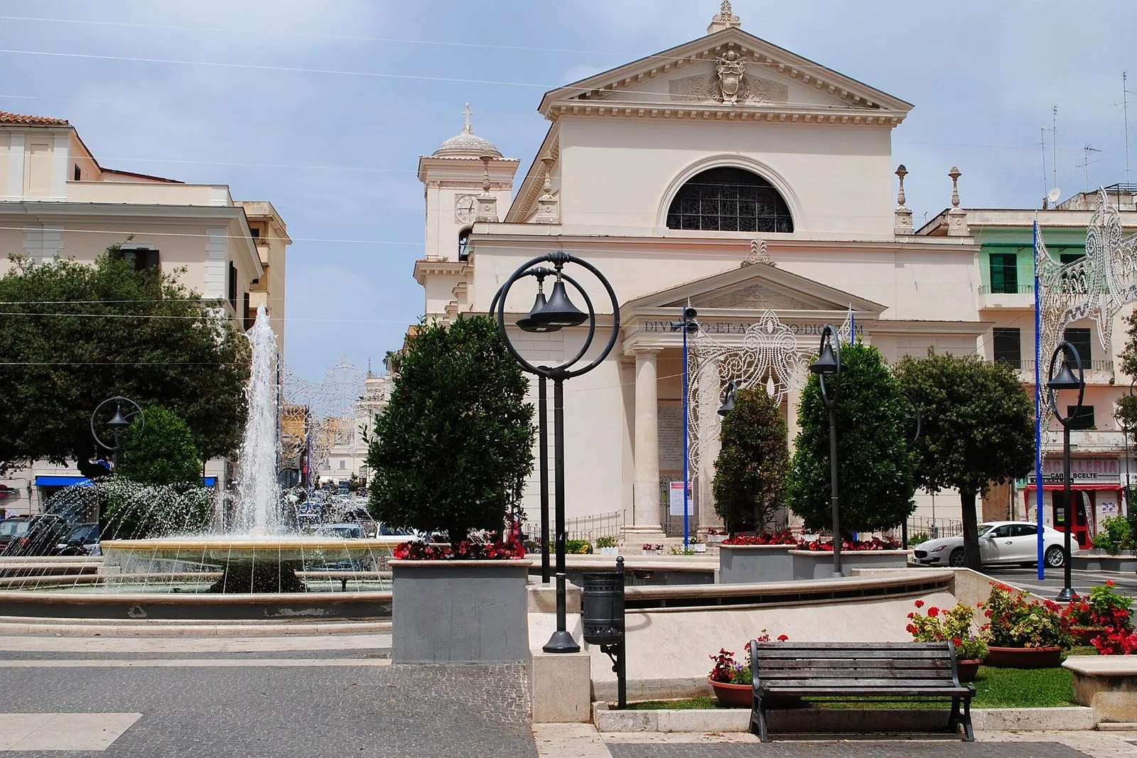 La facciata neoclassica della chiesa dei Santi Pio e Antonio ad Anzio, con il campanile e le luminarie di festa, vista dalla piazza con la fontana, gli alberi e le aiuole di gerani rossi