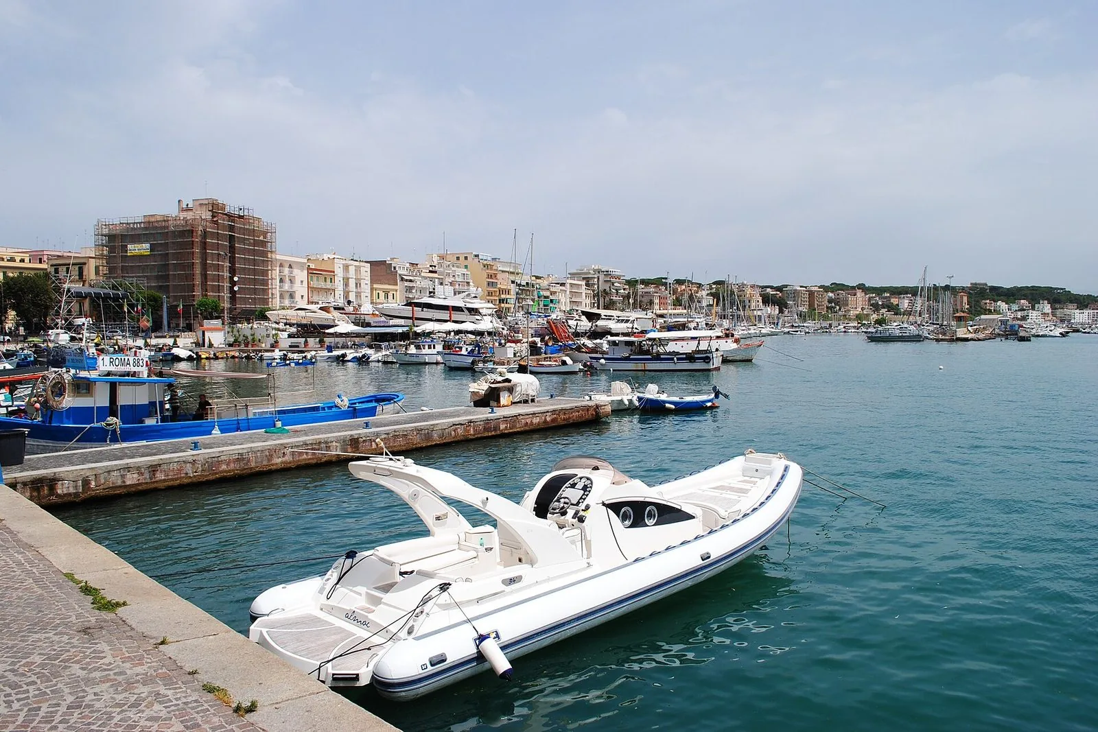 Il porto di Anzio visto dall'acqua: barche da pesca e yacht ormeggiati davanti a una fila di palazzi chiari e color ocra, con alberi di pino sulla collina e cielo grigio