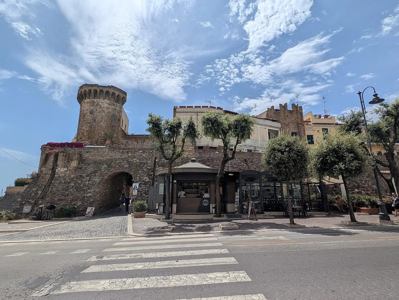 Le mura in pietra del borgo medievale di Nettuno con una grande torre cilindrica merlata, l'arco d'ingresso e alcuni alberelli lungo il viale, sotto un cielo azzurro con nuvole sottili