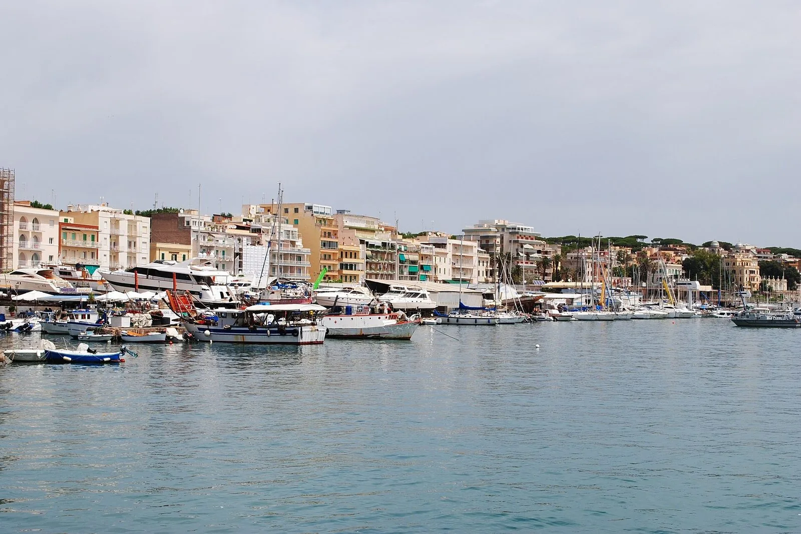 Il bacino del porto di Anzio sotto un cielo nuvoloso, con pescherecci, barche a vela e motoscafi ormeggiati davanti alla fila di palazzi e ai pini sulla collina