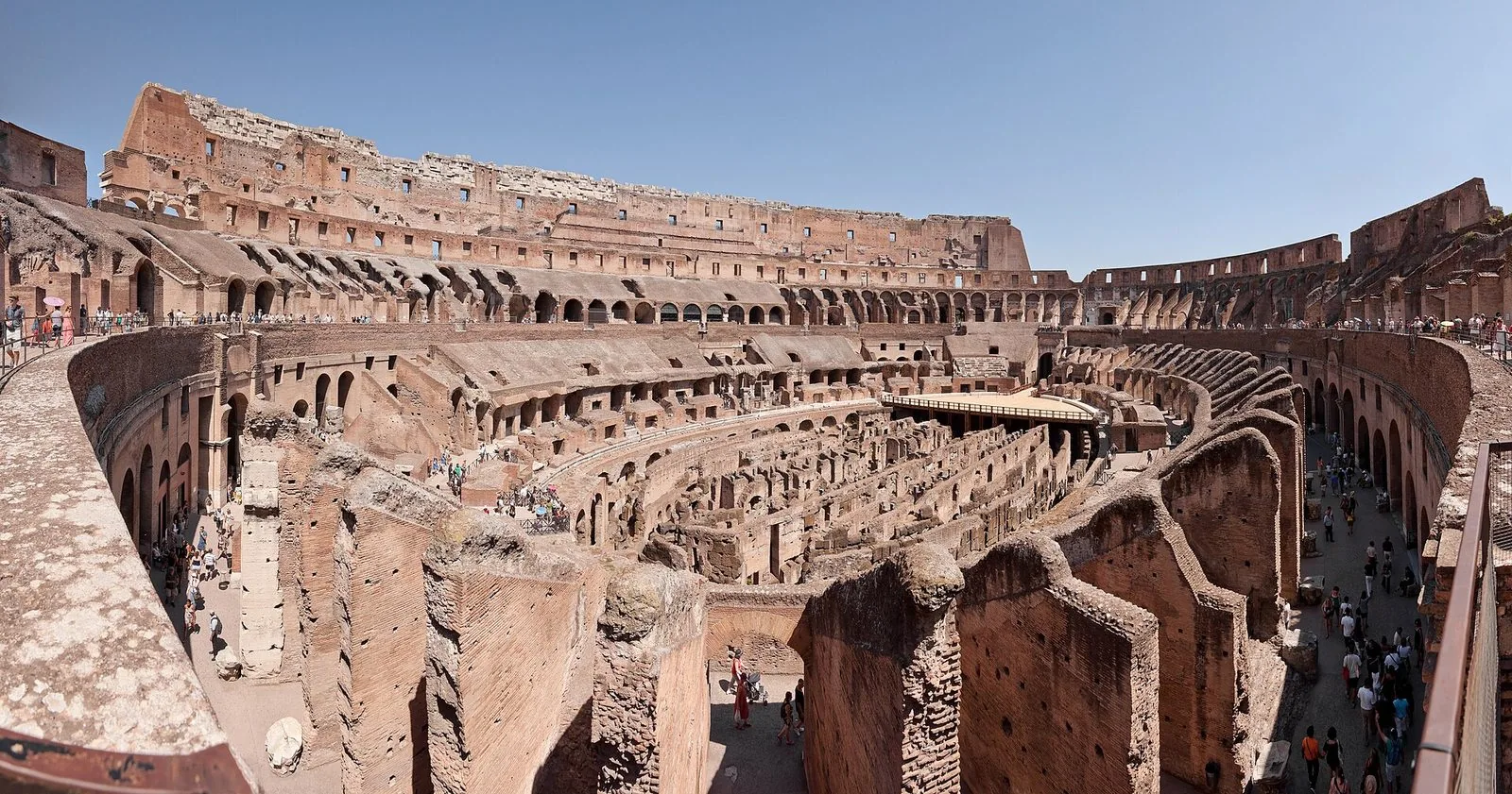 Veduta panoramica dell'interno del Colosseo: le gradinate in rovina, le arcate in travertino e mattoni e, al centro, i sotterranei dell'arena a vista con una piccola piattaforma in legno, sotto un cielo sereno