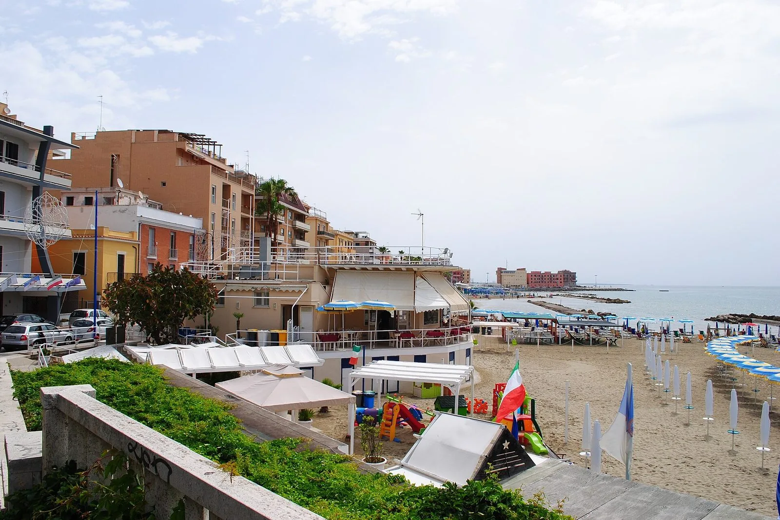La spiaggia di Anzio vista dal lungomare: ombrelloni chiusi allineati sulla sabbia, uno stabilimento con tende bianche e giochi per bambini, i palazzi della costa e il mare calmo con le scogliere frangiflutti